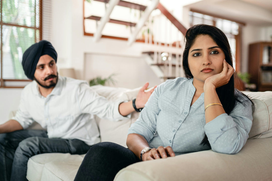 A married couple sits on a couch. The woman faces away, looking emotionally drained, while her husband tries to get her attention.