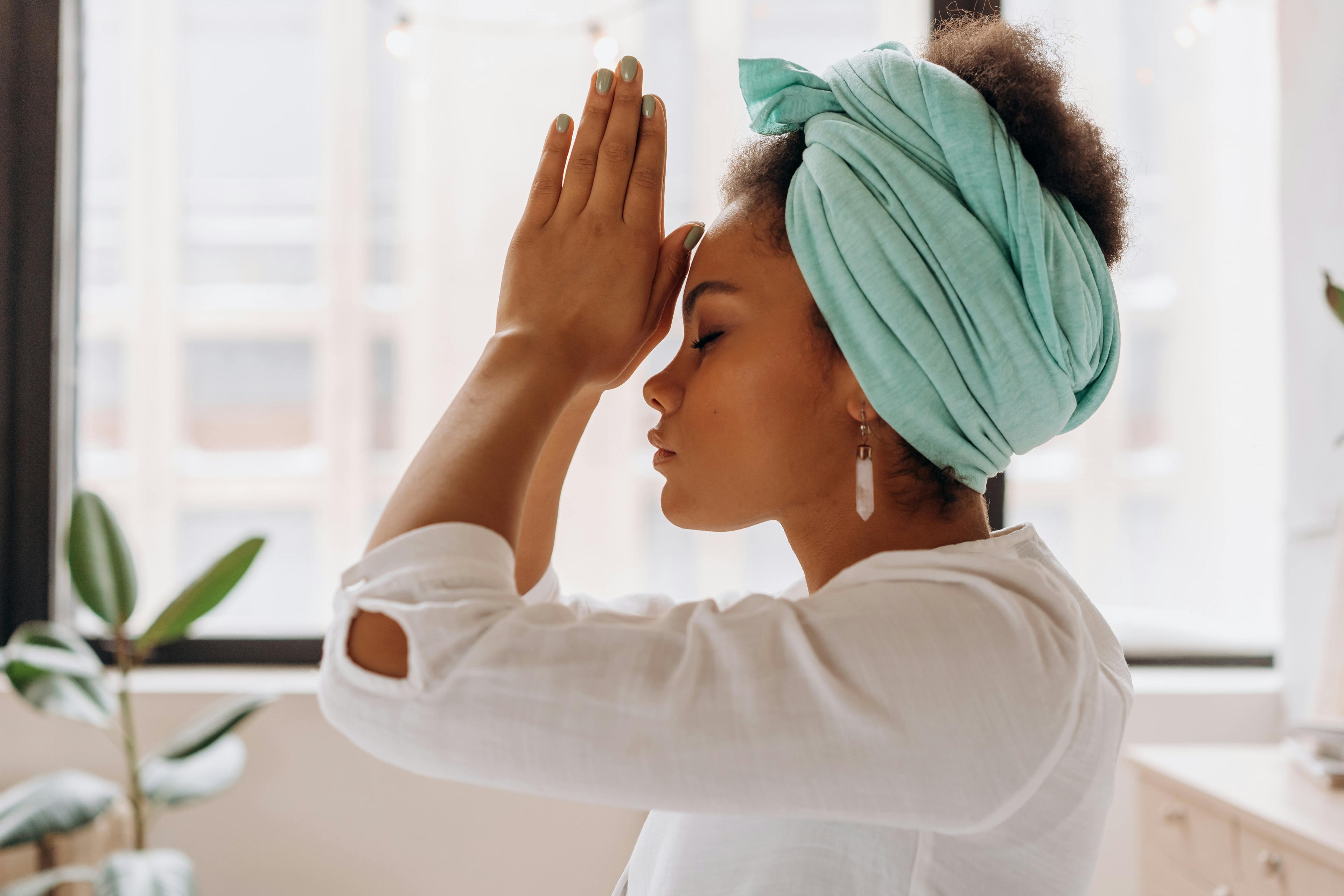 A young woman kneeling in her apartment with prayer hands in front of her face with her eyes closed