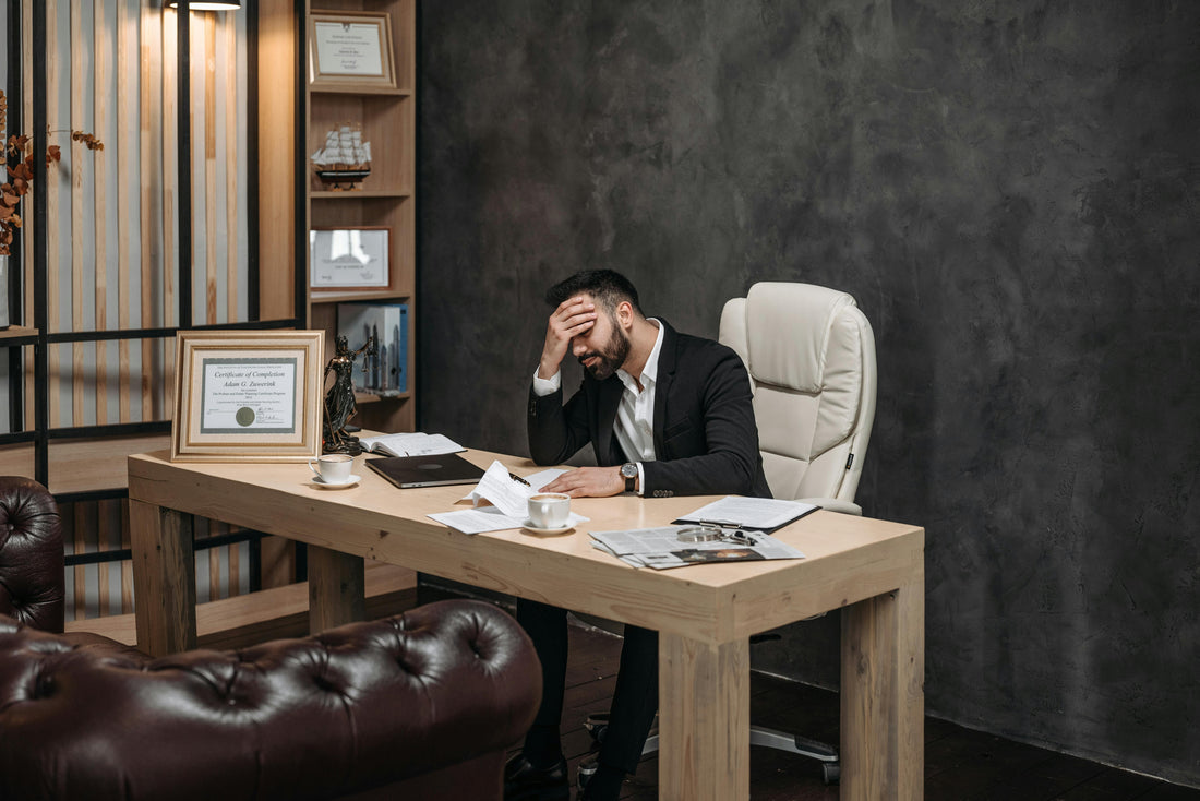 A man sitting at his office desk with his hand over his face, showing frustration and self-doubt, symbolizing imposter syndrome at work.