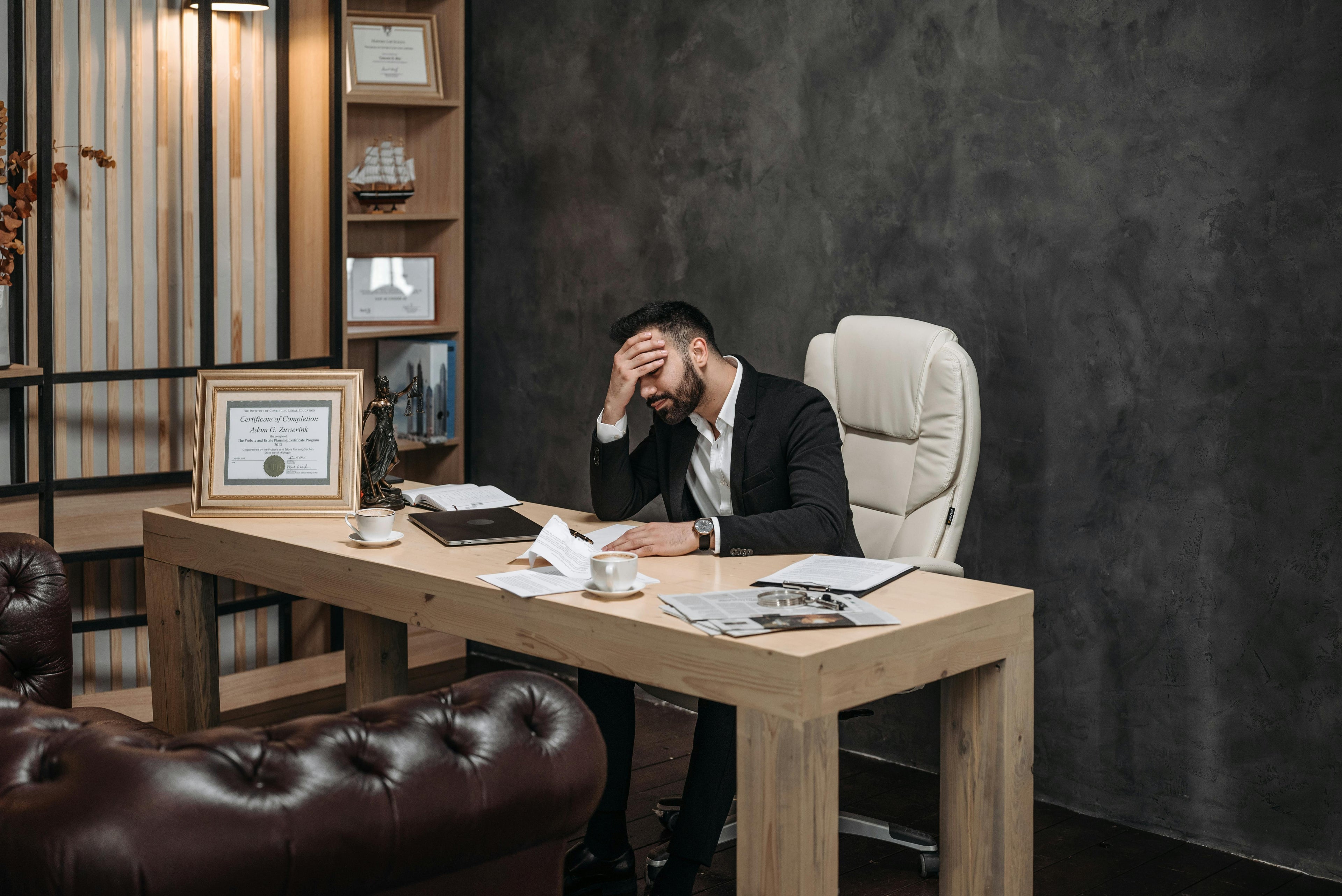 A man sitting at his office desk with his hand over his face, showing frustration and self-doubt, symbolizing imposter syndrome at work.