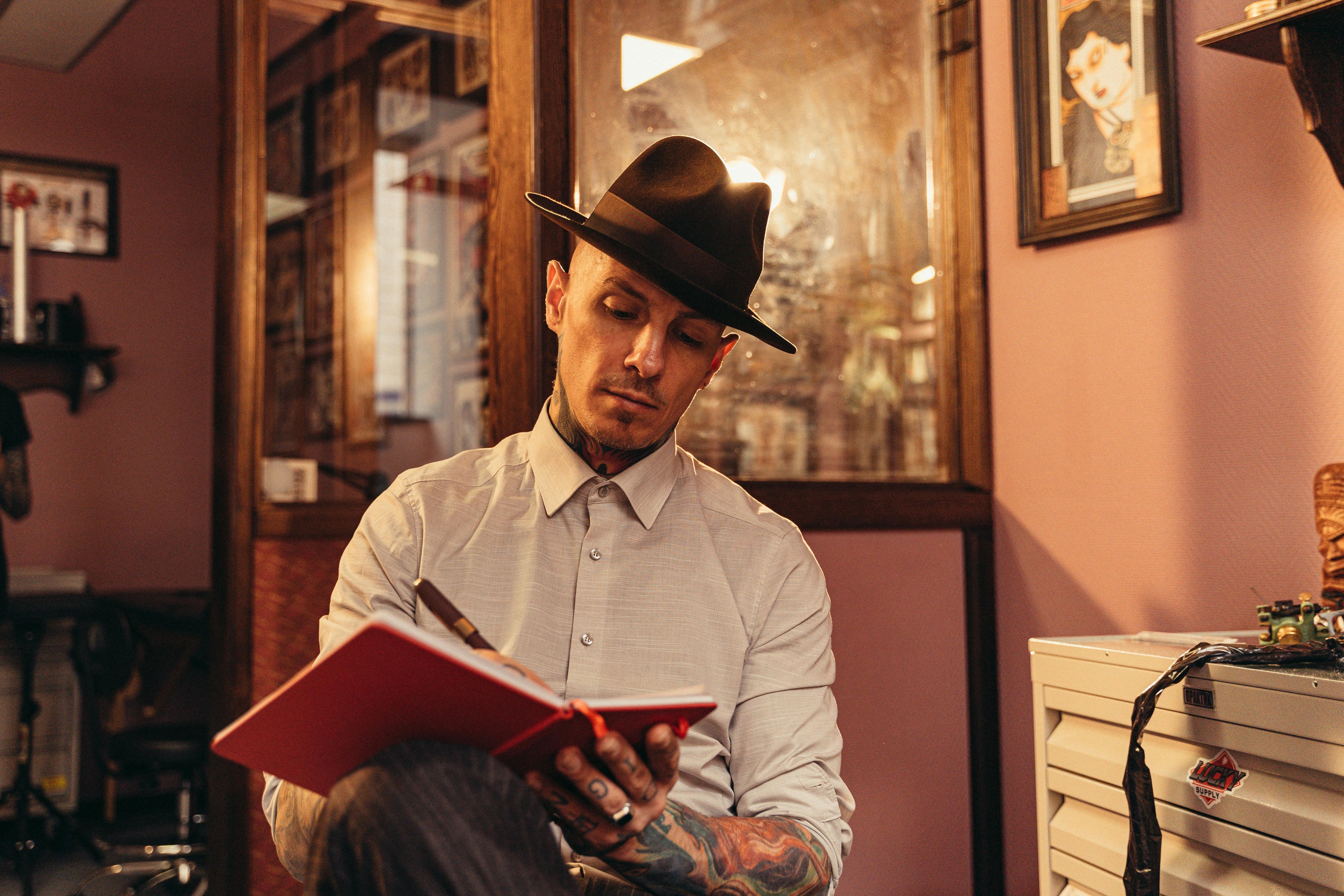 A thoughtful young man in a white cotton button-down shirt and fedora is seated and journaling, reflecting deeply as he writes in his notebook.