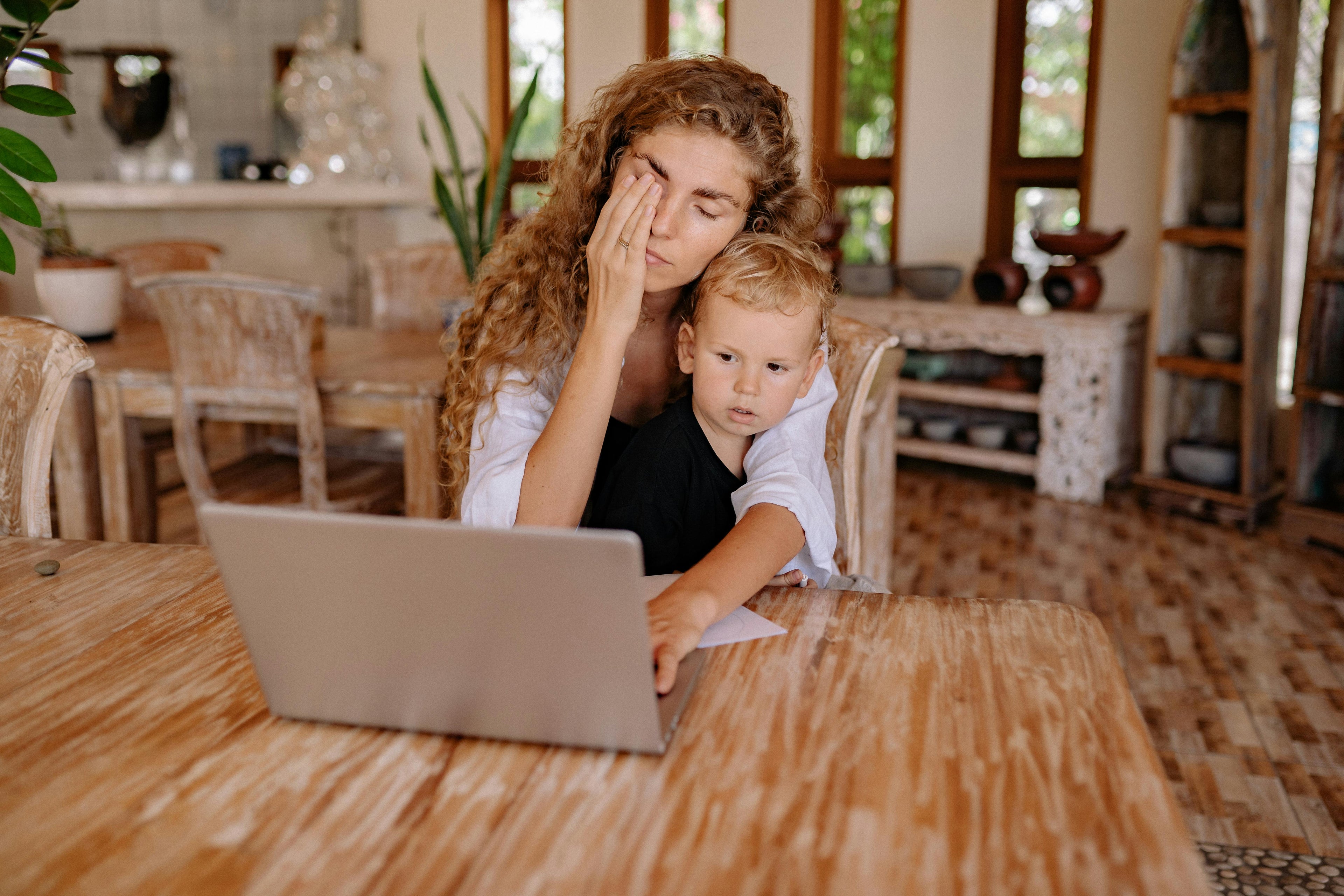 A tired young mother wearing a robe sits down, rubbing her eyes, with her small son on her lap. She looks emotionally and physically drained.